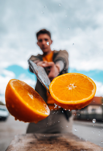 Close-up of a young man cutting a juicy orange in half with a large knife in motion, with splashes against the blue sky.