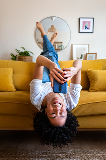 Feliz mujer afroamericana acostada boca abajo en el sofá enviando mensajes de texto usando el teléfono móvil. Revisando las redes sociales. Vertical. photo