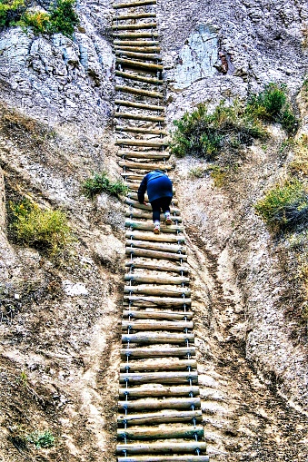 A lone individual, seen from the back at a distance, navigates a wooden ladder that climbs up a rocky cliff along a hiking trail in the Badlands of South Dakota.