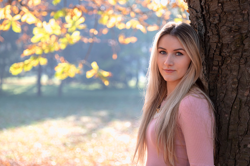 Close up portrait of attractive young woman in autumn park. She standing and leaning against a large tree and looks into the camera. She is wearing a pink sweater.