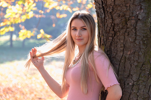 Close up portrait of attractive young woman in autumn park. She standing and leaning against a large tree and looks into the camera. She is wearing a pink sweater and playing with her long blonde hair.