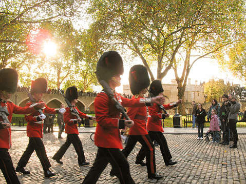 London, United Kingdom - October 26 2021: The British Royal Guard in red coats and black bearskin hats marching at sunset through the tower of London grounds with blurred motion and the sun shining through the trees.