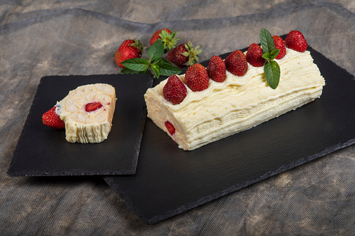 Sponge roll filled with cream and strawberries. Decorated with whipped cream. It lies on a slate tray ready for portioning and eating.