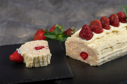 Sponge roll filled with cream and strawberries. Decorated with whipped cream. It lies on a slate tray ready for portioning and eating.