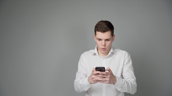 Surprised young guy in a white shirt looks at the screen of his smartphone. Grey background.
