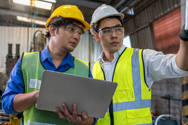 Foreman or Professional technician holding a laptop computer in an industrial factory. Male engineer apprentice trainee with machine in industrial machinery. stock photo