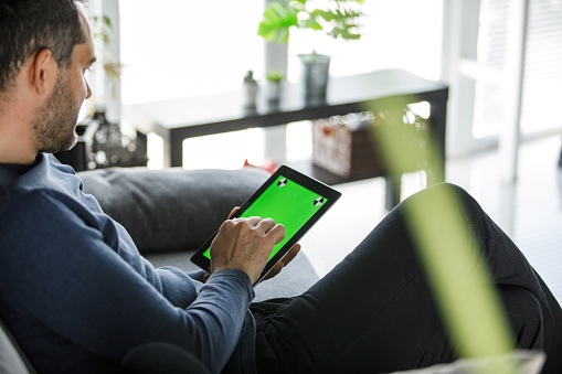 Three quarter length shot of contented mid adult man relaxing on the sofa and passing time by scrolling on digital tablet with chroma key on the screen.