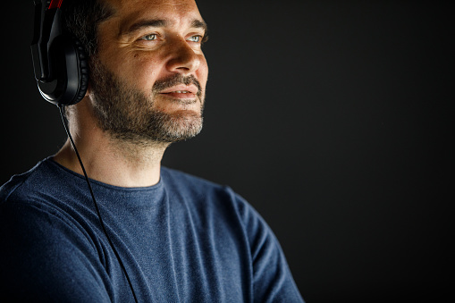 Copy space shot of smiling mid adult man standing against black background, listening to music via headphones, looking away, daydreaming.