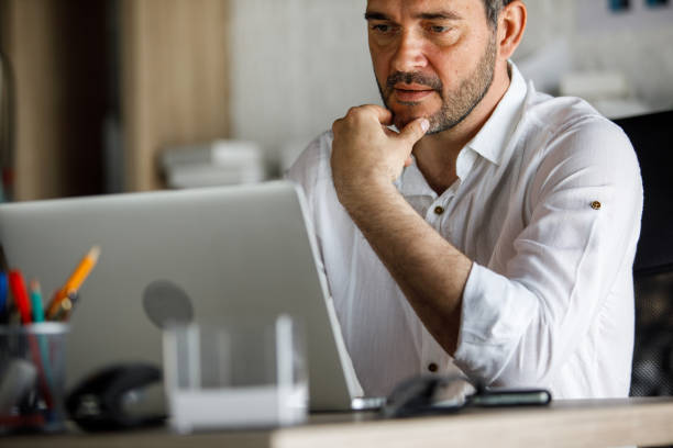 Copy space shot of focused mid adult businessman sitting at his desk at the office, hand under chin, analyzing data on laptop regarding a project he is working on.