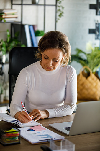 Portrait of diligent mature businesswoman sitting at her desk at the office, writing a to do list in her personal organizer while working.