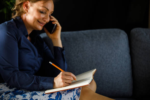 Selective focus shot of happy mature woman lounging on the sofa, talking on the phone with a friend and writing down plans in a notebook.