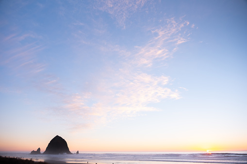 Oregon's Iconic Cannon Beach at sunset. Haystack Rock rises out of the sandy surf as cotton candy clouds float weightlessly in the orange pink and blue sky and the sun sets just over the horizon of the Pacific Ocean.