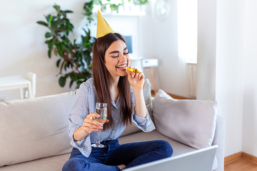 Virtual Celebration. Happy woman in party hat celebrating birthday online at quarantine or self-isolation, using laptop for video call with friends and family, holding champagne