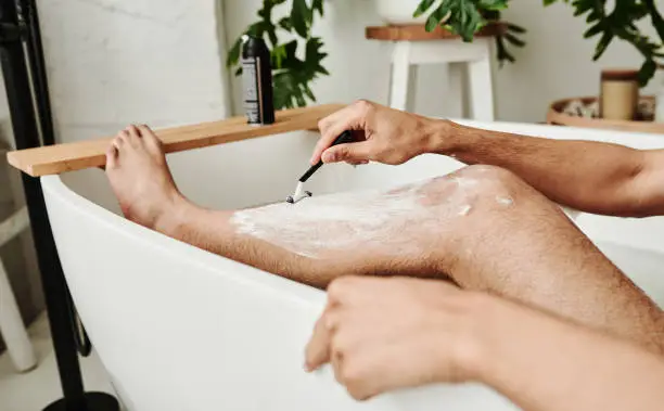 Close-up of young man using razor to shave his legs while sitting in bath