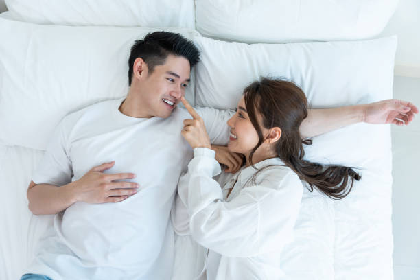 Portrait of Asian new marriage couple lying on bed and look each other. Attractive beautiful young man and woman in pajamas enjoy early morning activity in bedroom at home. Family relationship concept stock photo