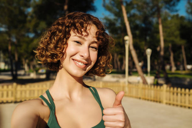 Sport selfie. Young attractive redhead smiling woman wearing green sport bra taking photo of herself before starting exercises, thumbs up, copy space. Green park background. Emotions, expressions stock photo