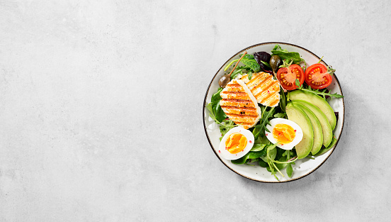 Healthy keto diet breakfast: boiled egg, avocado slices, grilled halloumi cheese, salad leaves. Light gray background. top view