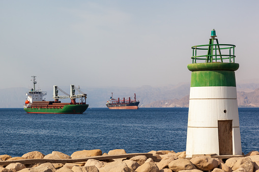 Small lighthouse on the coast of Aqaba, Jordan, with cargo ships in the background. Situated on the Red Sea, this busy port is the only sea port in Jordan