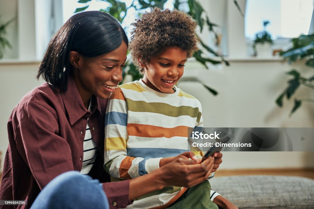 Boy and mom having fun while using phone at home Adorable little afro boy with curly hair and his mother using cellphone at home Child Stock Photo