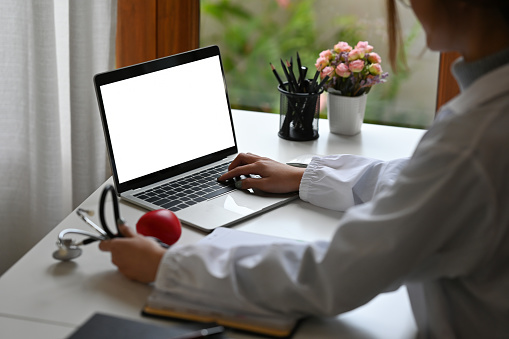 A cropped view of a young Asian doctor in white coat sitting in the office looking at the laptop and holding a stethoscope for medical and health care concept.