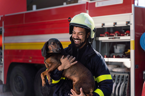 A happy mature firefighter man holding dog and looking at camera with fire truck in background