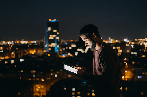 A young African American man standing on balcony with urban view and using tablet at night