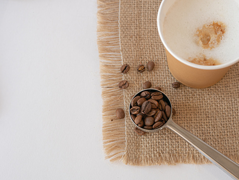 A disposable paper cup with cappuccino on a canvas napkin, grains and ground coffee are scattered on the table and a serving spoon. View from above. Space for copy.