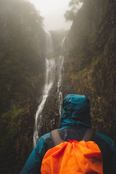 Rainy weather hiker in a waterproof jacket looks at the majestic Risco waterfall immersed in mist and rain on the island of Madeira, Portugal. Discovering magical places in Europe stock photo