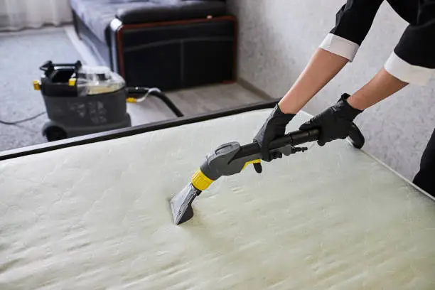 Cleaning service company employee removing dirt from furniture in flat with professional equipment. Female housekeeper arm cleaning the mattress on the bed with washing vacuum cleaner close up