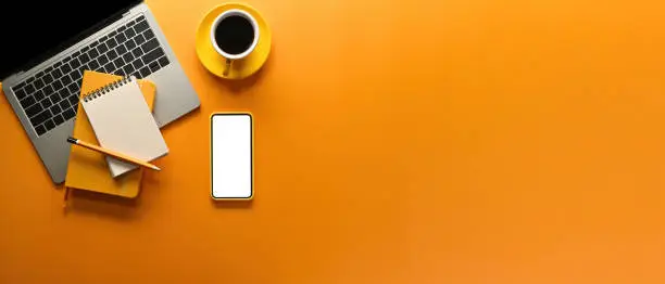 A top view of office flat lays such as note books, laptop, smart phone and a cup of coffee on an empty yellow-orange space on a background, for business and technology concept.