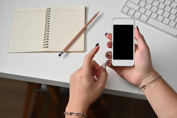 istock Top view of hands holding a smart phone with a black screen leaning on the white table with a note book on the left side, for business and technology concept. 1394602920