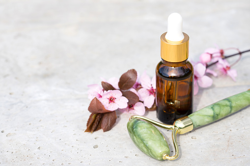 Facial massage kit on stone concrete background with copy space - jade gua sha face roller with beauty face serum or cosmetic and blooming sakura branch. Natural stone face massager, mockup