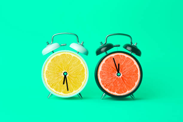 istock Alarm clock with citrus fruit concept. Lemon or orange and clock on empty color background. Vitamins, proper nutrition, health, detox, diet concept. 1394620525