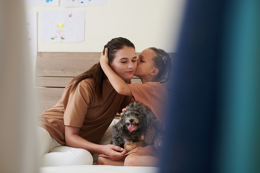 Girl kissing mother on cheek when they sitting on bed with small dog