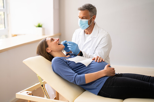 Male Dentist Treating Teeth Of Young Pregnant Woman Patient Lying In Clinic