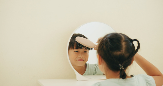 Asian little girl brushing her bangs hair with comb and smiling in the mirror of dressing table.