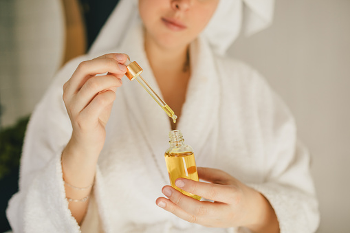 Beautiful young woman wearing white bathrobe and towel on head holding face oil in bathroom. Beauty cosmetic concept.