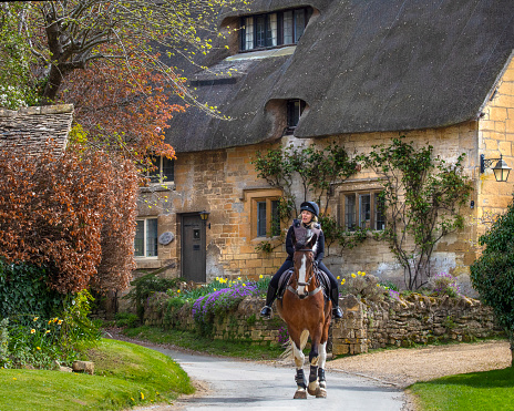 Gloucestershire, UK - April 11th 2022: A horse rider with a beautiful thatched cottage in the Cotswold village of Stanton in Gloucestershire, UK.