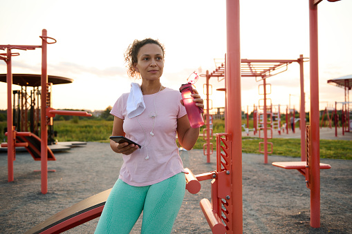 Beautiful Hispanic athletic woman resting on a sports field after workout on stationary gym machines and crossbar, holding a bottle with water and smartphone and looking away against sunset background