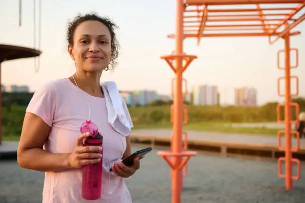 African American sportswoman with bottle of water and smartphone, smiles looking at camera standing on a sports field, working out at sunset