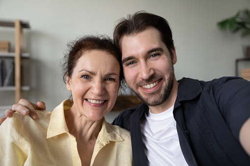 Happy adult son and mature mom hugging with love, affection, taking selfie, looking, smiling at camera, having fun, enjoying leisure at home together. Head shot portrait. Motherhood concept