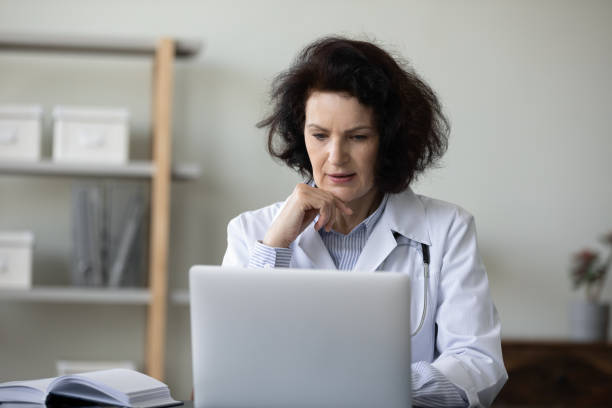 serious focused senior doctor working at laptop in hospital office - huisarts druk stockfoto's en -beelden