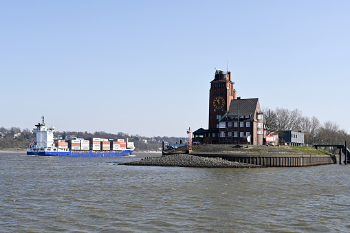 Hamburg, Germany, March 20, 2022 - Pilot house Seemannslöft in Hamburg Finkenwerder and a container ship  Katharina Schepers on the river Elbe
