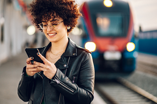 Young businesswoman with afro hair standing on railway station platform and using a smartphone while waiting for a train. Checking message sms e-mail or train schedule.