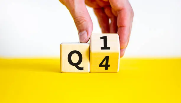 Photo of From 4th to 1st quarter symbol. Businessman turns a wooden cube and changes words 'Q4' to 'Q1'. Beautiful yellow table, white background. Business, happy 1st quarter Q1 concept, copy space.