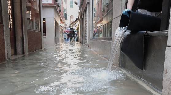 person empties the bucket of water from inside the flooded shop in the alley of Venice in Italy during high tide