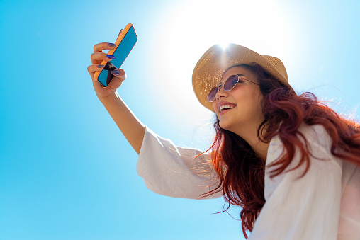 Smiling pretty young girl taking selfie on the beach. Sunglasses, straw hat, pareo, cell phone, red hair.