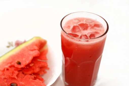 A fresh watermelon smoothie juice drink with ice cubes and a plate full of Slices of watermelon fruit cuts into pieces isolated on a white background, summer tropical refreshments and drinks concept