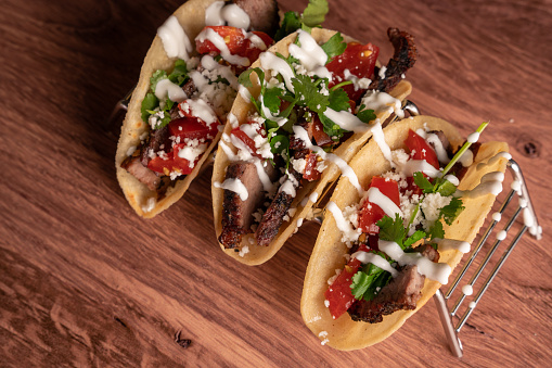 Gourmet Carne Asada Street Tacos lined on a metal serving rack with tomato salsa, sour cream and cilantro ready to eaten. Authentic Mexican Cuisine on a dark back ground with studio lighting to make the taco stand out.