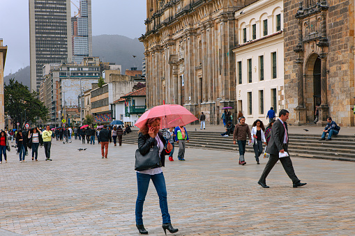 Bogota, Colombia - July 01, 2016: Local Colombian people are seen going about their day to day lives on Bolivar Square in the Andean Capital city of Bogota in Colombia, South America. Centre right is the 19 Century Catedral Primada and to its right is the entrance to the 18th Century Baroque style Capilla del Sagrario. In the far background are the Andes Mountains. The sky is overcast. The altutude at street level is 8,660 feet above sea level. Most people wear warm clothes. Photo shot in the afternoon sunlight on an overcast day; horizontal format. Copy space.
